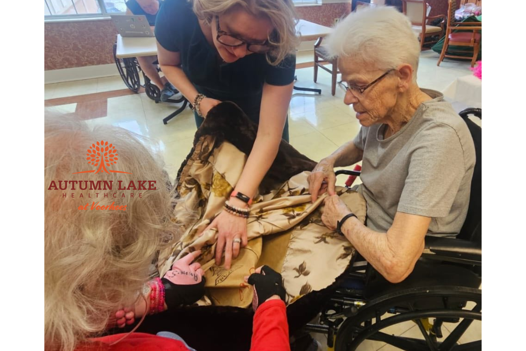 A staff member and two senior living residents in wheelchairs work together on a patterned fabric craft project in a sunlit