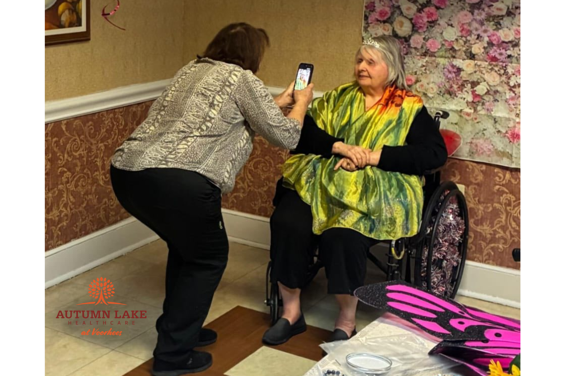 A staff member takes a photo of an assisted living resident dressed in a colorful shawl and tiara during a glamorous dress-up event.