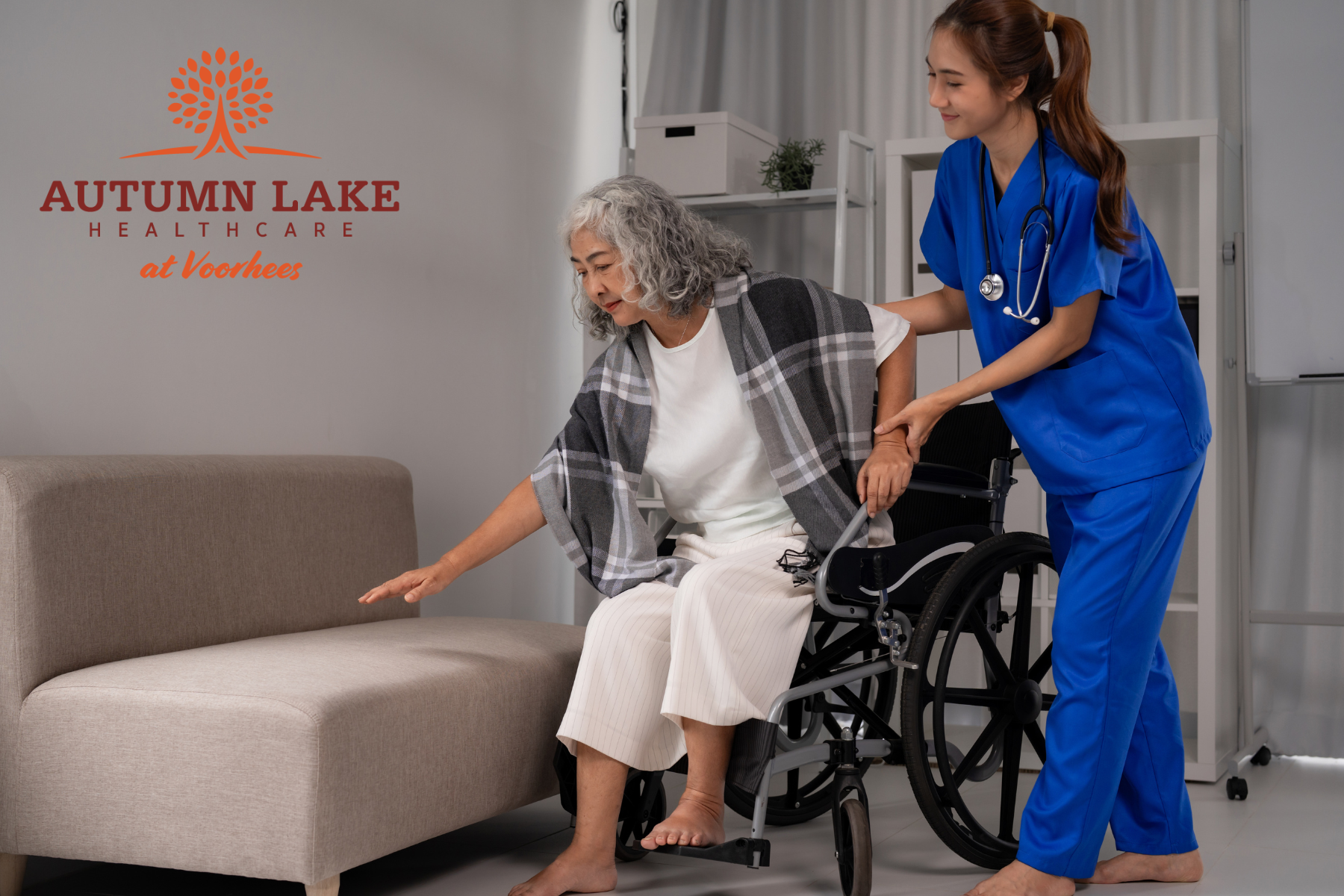 Nurse assisting a senior resident transitioning from a wheelchair to a sofa in an assisted living facility.